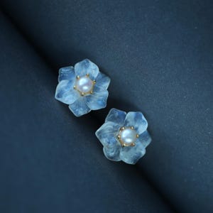 Könnte beinhalten: Ein Paar blütenförmige Ohrringe. Jeder Ohrring hat ein hellblaues, durchscheinendes Blumendesign mit einer zentralen Perle und goldfarbenen Akzenten. Die Ohrringe sind vor einem dunkelblauen Hintergrund platziert.