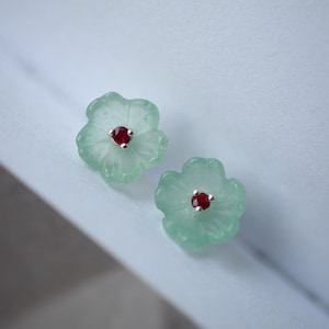 Könnte beinhalten: Ein Paar blütenförmige Ohrringe. Jeder Ohrring hat ein hellgrünes, geschnitztes Blumenmuster mit einem kleinen, runden, roten Edelstein in der Mitte. Die Ohrringe sind auf einem Silberstift befestigt.
