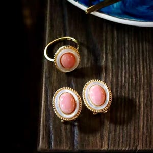 May include: A gold-coloured jewellery set comprising a ring and two earrings. Each piece features a central oval pink stone, framed by a white border and a gold-coloured rope-like trim. The set is presented on a dark wooden surface.