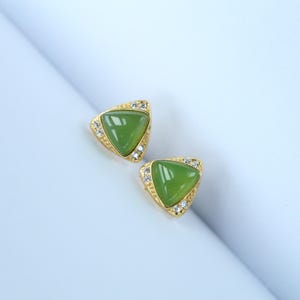 Puede incluir: Un par de pendientes de botón dorados. Cada pendiente presenta una piedra triangular verde claro rodeada de pequeños cristales transparentes. Los pendientes están sobre un fondo blanco y azul claro.