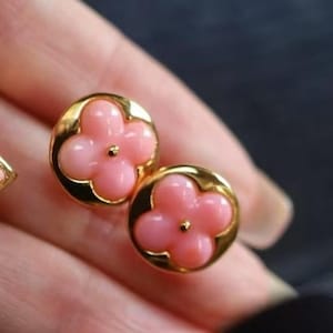 Può includere: Un paio di orecchini a bottone dorati. Ogni orecchino presenta un design a fiore rosa a quattro petali incastonato in una cornice circolare dorata. Gli orecchini sono piccoli e delicati, con una finitura lucida.