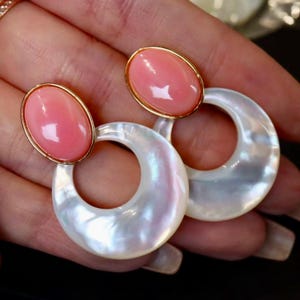 Può includere: Orecchini con una gemma rosa ovale incastonata in una montatura dorata, collegata a un grande elemento bianco iridescente a forma di mezzaluna. Gli orecchini sono presentati su uno sfondo scuro.