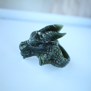 Puede incluir: Un anillo de dragón verde oscuro tallado en piedra. El anillo presenta una cabeza de dragón detallada con escamas texturizadas, cuernos y dientes afilados. Una pieza de joyería única.