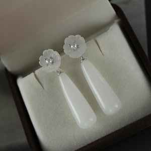 Può includere: Un paio di orecchini pendenti bianchi con un design floreale nella parte superiore e un pendente a forma di goccia. Gli orecchini sono esposti in un portagioie marrone con interno color crema. Gli orecchini hanno un gancio in metallo argentato.