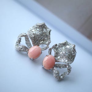 Può includere: Un paio di orecchini argentati con un design a farfalla. Ogni orecchino presenta una grande pietra ovale rosa chiaro al centro, circondata da pietre preziose più piccole e trasparenti. Le ali sono strutturate e dettagliate.