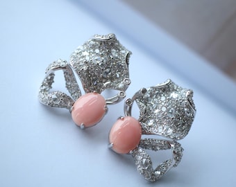 May include: A pair of silver-toned earrings with a butterfly design. Each earring features a large, oval, light pink gemstone at the center, surrounded by a cluster of smaller, clear gemstones. The wings are textured and detailed.