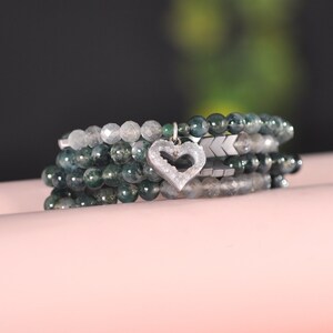 Könnte beinhalten: Ein Set aus vier grünen und grauen Agat-Armbändern mit einem silbernen Herz-Anhänger. Die Armbänder sind übereinander gestapelt und liegen auf einer rosa Oberfläche.