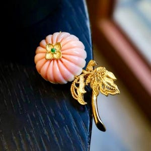 Peut inclure: Une broche en or ornée d'une pierre sculptée rose pâle en forme de citrouille, surmontée d'une petite fleur en or texturé avec une minuscule pierre verte au centre. Elle comprend une tige dorée et deux feuilles texturées détaillées.
