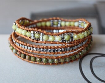 May include: A multi-strand leather wrap bracelet with green and silver beads. The bracelet is made with brown leather cord and features green gemstone beads and silver square beads.
