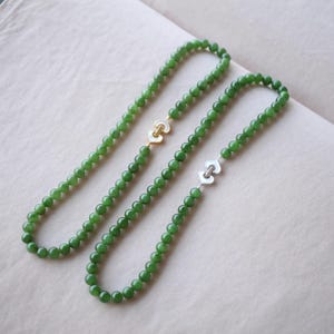 May include: Three green jade bead necklaces. Two necklaces have heart-shaped clasps, one gold-toned and the other silver-toned. The necklaces are displayed on a neutral-colored surface.