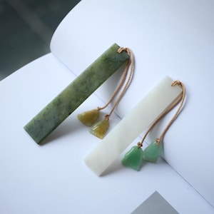 Puede incluir: Dos marcapáginas rectangulares de jade. Uno es verde moteado, el otro blanco. Cada uno tiene una borla de pequeños amuletos de jade en tonos amarillos y verdes, sujetos con un fino cordón marrón. Los marcapáginas están sobre una superficie blanca.