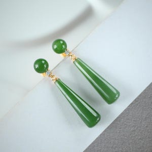 Può includere: Un paio di eleganti orecchini di giada verde. Ogni orecchino presenta un perno rotondo di giada verde, un piccolo accento di diamante e un lungo pendente di giada verde a forma di goccia. Gli orecchini hanno dettagli in metallo color oro.