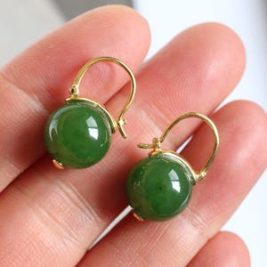 Op de afbeelding: Een paar goudkleurige oorringen, elk met een ronde, gepolijste, diepgroene jade steen. De oorbellen worden in een hand gehouden, wat het contrast tussen het goudmetaal en de levendige groene edelstenen benadrukt. De oorbellen zijn een klassiek sieradenontwerp.