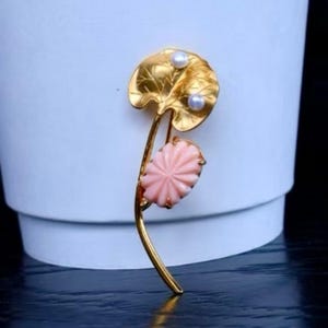 Peut inclure: Broche dorée en forme de fleur. La broche présente une feuille d'or détaillée, deux petites perles et une fleur rose sculptée. La tige est incurvée, le tout sur fond blanc.