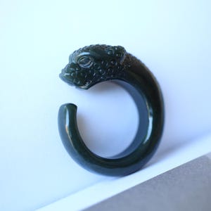 May include: A dark green jade ring carved in the shape of a jaguar. The ring is smooth and polished, with intricate details on the jaguar's face and body.