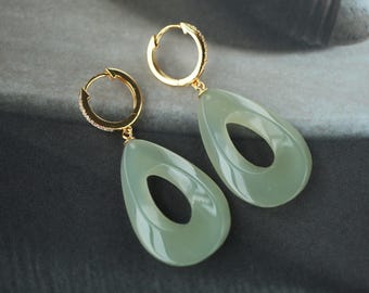 May include: A pair of gold-colored hoop earrings with a light green, teardrop-shaped pendant. The pendants have a large oval cutout in the center. The earrings are displayed on a dark gray surface.