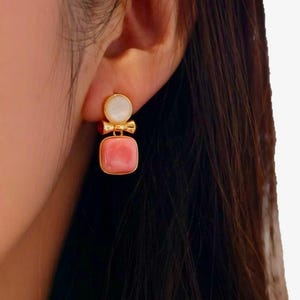 Può includere: Orecchini dorati con un design a due pietre. La pietra superiore è bianca e circolare, mentre la pietra inferiore è una gemma rosa quadrata. Un piccolo fiocco dorato collega le due pietre. Gli orecchini sono indossati su un orecchio.