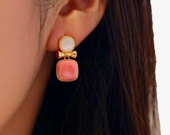 May include: Gold-toned earrings with a two-stone design. The top stone is a white, circular shape, and the bottom stone is a square, pink gemstone. A small gold bow connects the two stones. The earrings are worn on a person's ear.