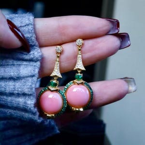 Può includere: Orecchini color oro con un grande centro di pietre preziose rosa rotonde, circondato da piccole pietre verdi. Gli orecchini hanno una parte superiore decorativa con pietre chiare e un piccolo accento di pietra verde.