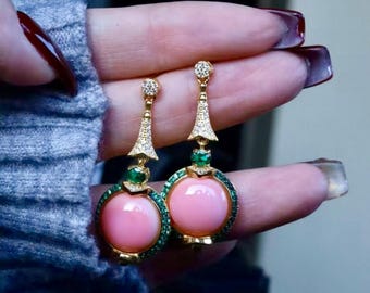 May include: A pair of gold-toned earrings featuring a large, round, pink gemstone center, surrounded by a ring of small green stones. The earrings have a decorative top with clear stones and a small green stone accent.