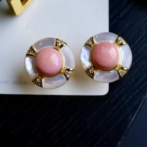 Può includere: Un paio di orecchini a bottone rotondi. Ogni orecchino presenta una pietra centrale rosa chiaro, incorniciata in oro e un anello esterno bianco iridescente. Piccole pietre trasparenti sono incastonate nella montatura dorata.