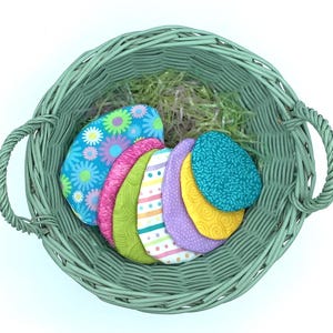 Peut inclure: Un panier en osier avec une finition vert lavé, rempli d'œufs de Pâques en tissu colorés. Les œufs sont de différents motifs, notamment floraux, rayés et à pois.