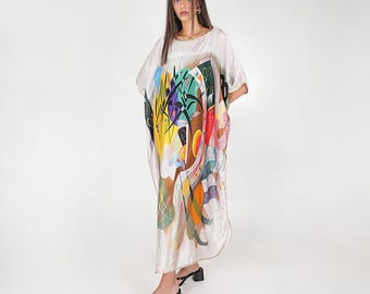 May include: A floor-length, loose-fitting kaftan dress in a light cream color, featuring a vibrant, abstract graphic print with bold shapes in yellow, red, blue, green, and black. The garment has short, wide sleeves and a round neckline.
