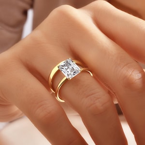 May include: A gold ring with a large, clear, rectangular-shaped gemstone. The ring has a double band design.