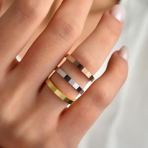 May include: Four rings in gold, silver, and rose gold are stacked on a finger. The rings have a flat, geometric design, with a square profile. The rings are displayed against a soft, neutral background.