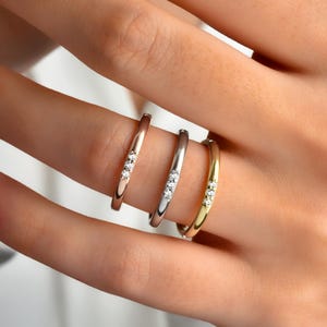 May include: Four rings in rose gold, silver, and gold, each with three small, clear gemstones. The rings are displayed on a finger, showcasing their different metallic finishes and sparkling details. The rings are delicate and elegant.