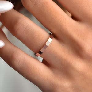 May include: A rose gold ring with a geometric design. The ring has a series of small, rectangular facets, creating a textured look. The ring is worn on a finger, showcasing its minimalist and modern style.