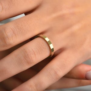 May include: A gold ring with a geometric design. The ring is worn on a finger and features small, rectangular facets around its band. The ring's surface reflects light, creating a bright, shiny appearance. The ring is a piece of jewellery.