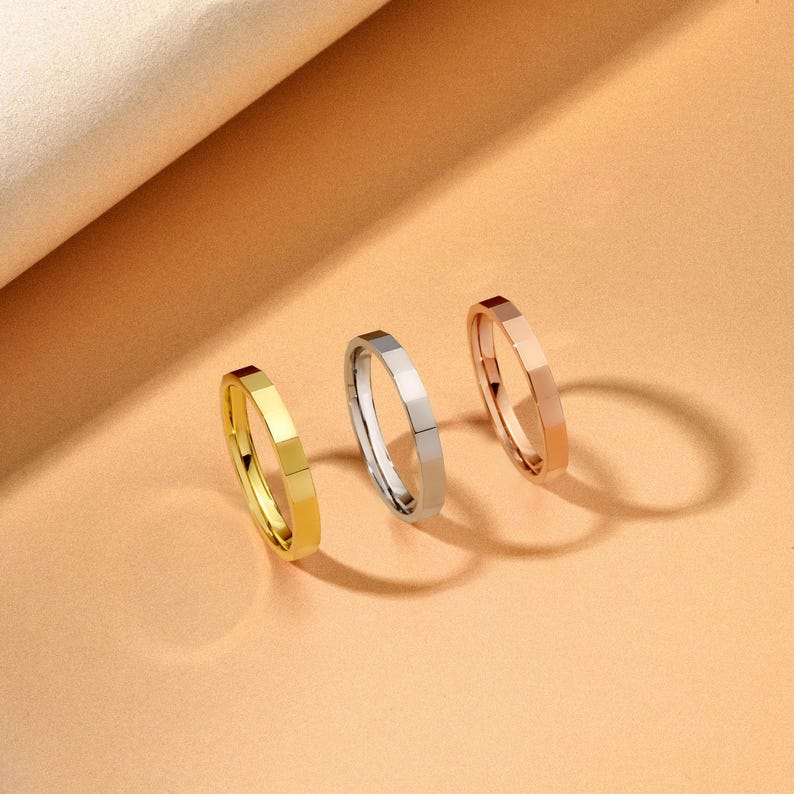 May include: Three minimalist rings in gold, silver, and rose gold. Each ring has a faceted design, creating a geometric look. The rings are displayed on a peach-coloured surface, with a soft, neutral background.