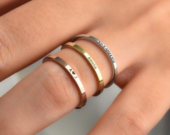 May include: A close-up of three thin, personalized rings in rose gold, gold, and silver. The rose gold ring has "A ♥ J" engraved. The gold ring is engraved with "Amelia". The silver ring has "XII-X-MMXXIV" engraved.