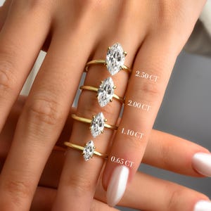 May include: Four gold rings with marquise-cut gemstones of varying sizes are displayed. The rings are labeled with their carat weights: 2.50 CT, 2.00 CT, 1.10 CT, and 0.65 CT. The rings are on a hand with manicured nails.