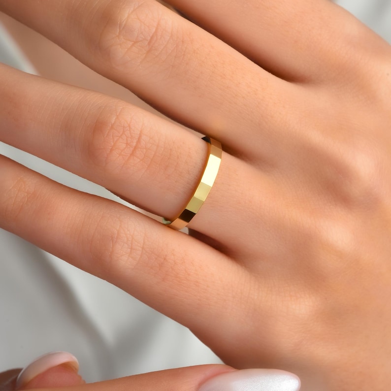 May include: A close-up of a gold ring with a geometric design. The ring is on a finger, and the gold colour is bright and shiny. The ring's surface is faceted, creating a textured appearance. The ring is a simple band style.