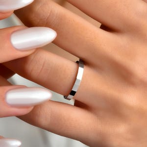 May include: A silver ring with a geometric design, featuring multiple rectangular facets. The ring is worn on a finger. The image shows a close-up of the ring and the hand, with white nail polish.