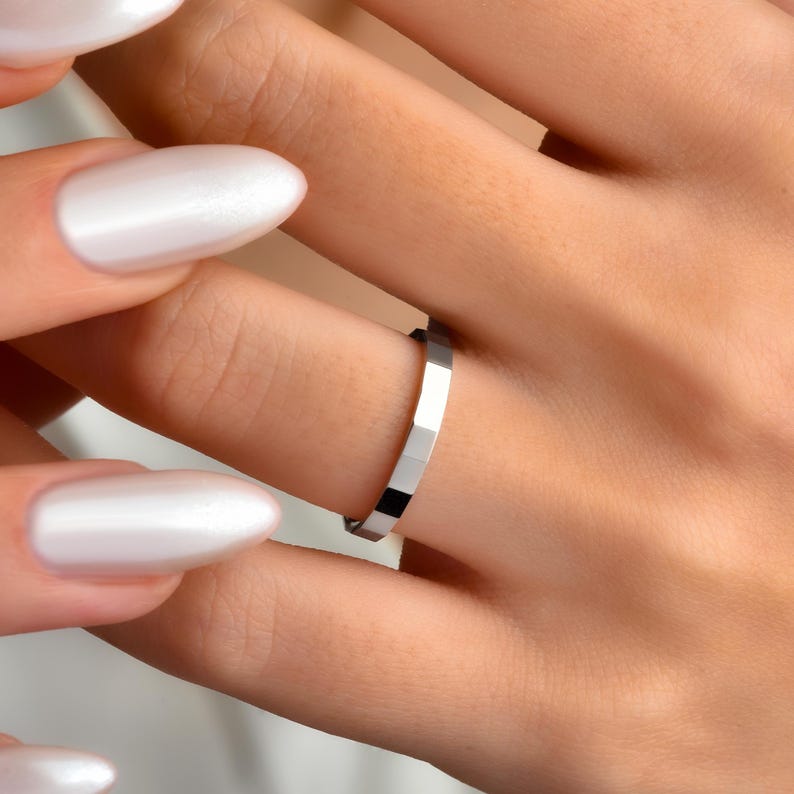 May include: A silver ring with a geometric design, featuring multiple rectangular facets. The ring is worn on a finger. The image shows a close-up of the ring and the hand, with white nail polish.