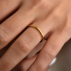 May include: A thin, gold-colored ring is worn on a finger. The ring has a simple, minimalist design, with a smooth, rounded surface. The ring appears to be made of a shiny metal, reflecting the light.