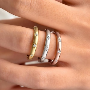 May include: Three rings in gold, silver, and rose gold, each adorned with star-shaped embellishments. The rings are displayed on a finger, showcasing their different metallic finishes and sparkling details. A close-up shot highlights the jewelry's design.