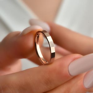 May include: A rose gold ring with a geometric, faceted design. The ring is held between two fingers, showcasing its polished surface and unique shape. The ring's design features a series of flat, angled facets that catch the light.