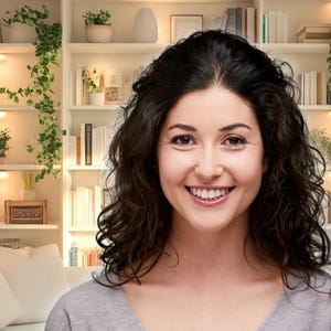 May include: A woman with long, dark curly hair smiles at the camera. She is wearing a light gray shirt and is standing in front of a bookshelf filled with books and plants.