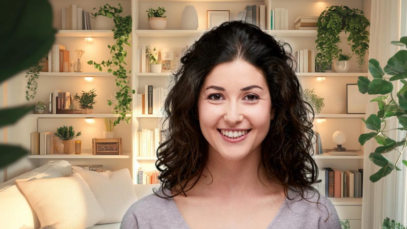 May include: A woman with long, dark curly hair smiles at the camera. She is wearing a light gray shirt and is standing in front of a bookshelf filled with books and plants.
