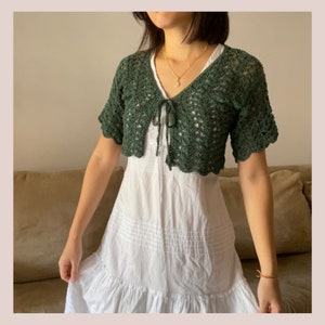 May include: A green crocheted cropped cardigan with a tie front. The cardigan has a delicate openwork pattern and short sleeves.