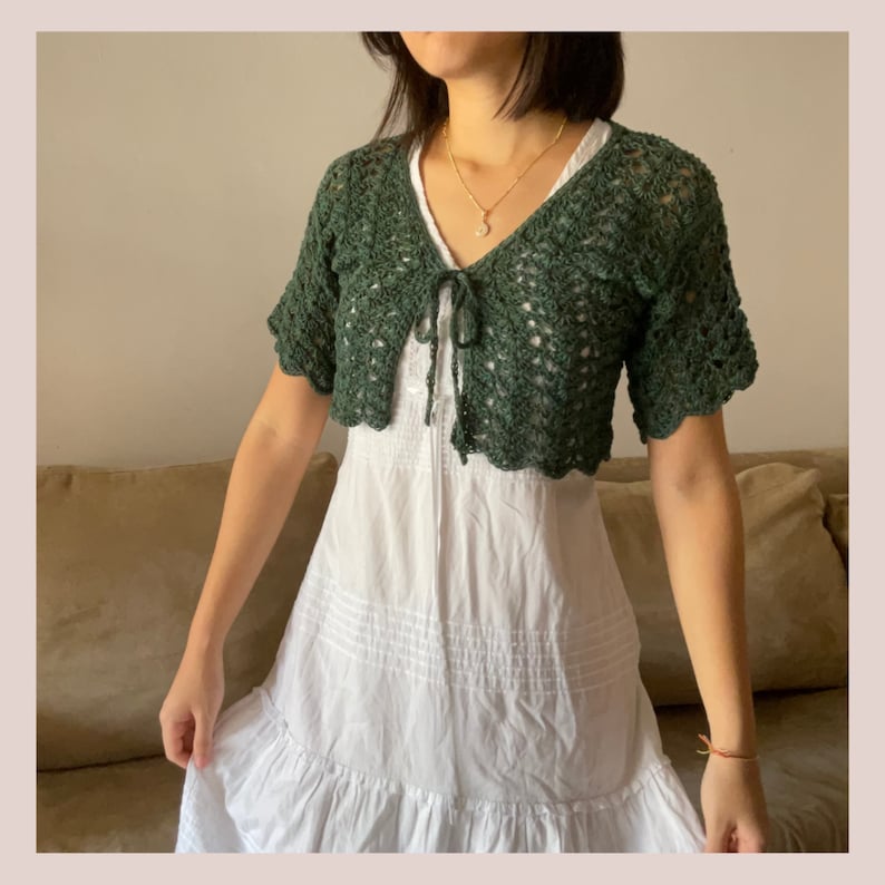 May include: A green crocheted cropped cardigan with a tie front. The cardigan has a delicate openwork pattern and short sleeves.