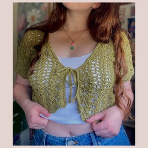May include: A green crocheted crop top with a tie front. The top is worn over a white tank top and blue jeans.