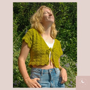 May include: A woman wearing a green crocheted crop top with a tie front. The top has a scalloped hem and short sleeves. She is wearing blue jeans and has her hair pulled back.