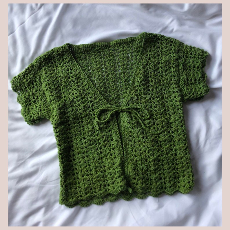 May include: A green crocheted cardigan with a tie front and short sleeves. The cardigan has a scalloped hem and is made with a delicate stitch.