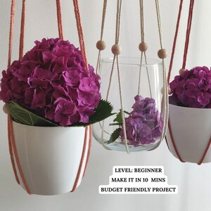 May include: Three hanging plant holders with pink and white macrame cord and wooden beads. Two holders have pink hydrangeas in white pots and one has a glass vase with purple hydrangeas. The text on the image reads "Level: Beginner, Make it in 10 mins, Budget Friendly Project."