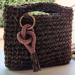 May include: A brown crocheted bag with a gold ring and a pink tassel. The bag is made of a soft, fuzzy material.